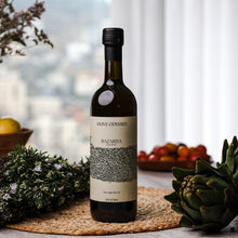 Load image into Gallery viewer, Bottle of Olive Odyssey Bazariya extra virgin olive oil on a woven mat with artichokes and fruit in the background.