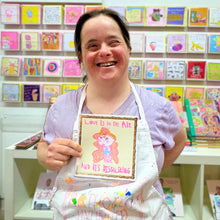 Load image into Gallery viewer, Arthouse Unlimited artist holding a card with 'Love is in the Air and it's Delicious' text in a store with colourful cards on shelves in the background