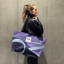 Load image into Gallery viewer, Female model wearing the large recycled tote bag in purple tones from the Upcycled by V&A Dundee collection.