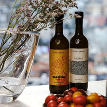 Load image into Gallery viewer, Two bottles of Olive Odyssey artisan Palestinian olive oil with a vase of flowers and fruits on a table.
