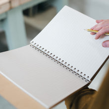 Load image into Gallery viewer, Open refillable walnut notebook with lined recycled coffee paper insert being used for writing.