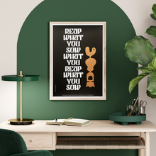 Load image into Gallery viewer, Framed graphic art work with words 'You Reap What You Sow' with a black print background, white text and a metallic motif. Art work set in an office space on a wall with bold green decorative arch paintwork and a table lamp, greenery in a modern office set up
