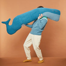 Load image into Gallery viewer, Man holding a large decorative blue knitted whale cushion set against an orange background
