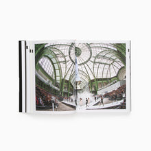 Load image into Gallery viewer, Open book with double page spread showing The Chanel "Centre de Lancement No 5" fashion show with an interior view of the grand building with a glass ceiling and the runway with models walking also the audience
