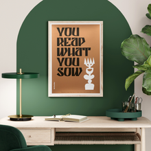 Load image into Gallery viewer, Framed graphic art work with words 'You Reap What You Sow' set in an office space on a wall with bold green decorative arch paintwork and a table lamp, greenery in a modern office set up
