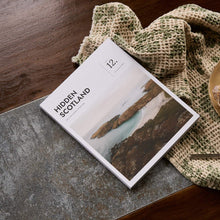 Load image into Gallery viewer,  'Hidden Scotland' magazine issue 12 Spring Summer 2026 sitting on a desk next to a plate of cake on a textured surface

