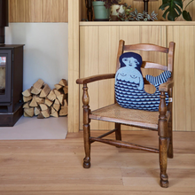 Load image into Gallery viewer, Blue knitted mermaid cushion on a wooden chair in a room setting with log burner in the background. 

