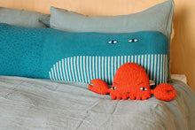 Load image into Gallery viewer, Whale bolster cushion by Donna Wilson sitting across a bed next to a small knitted crab cushion.
